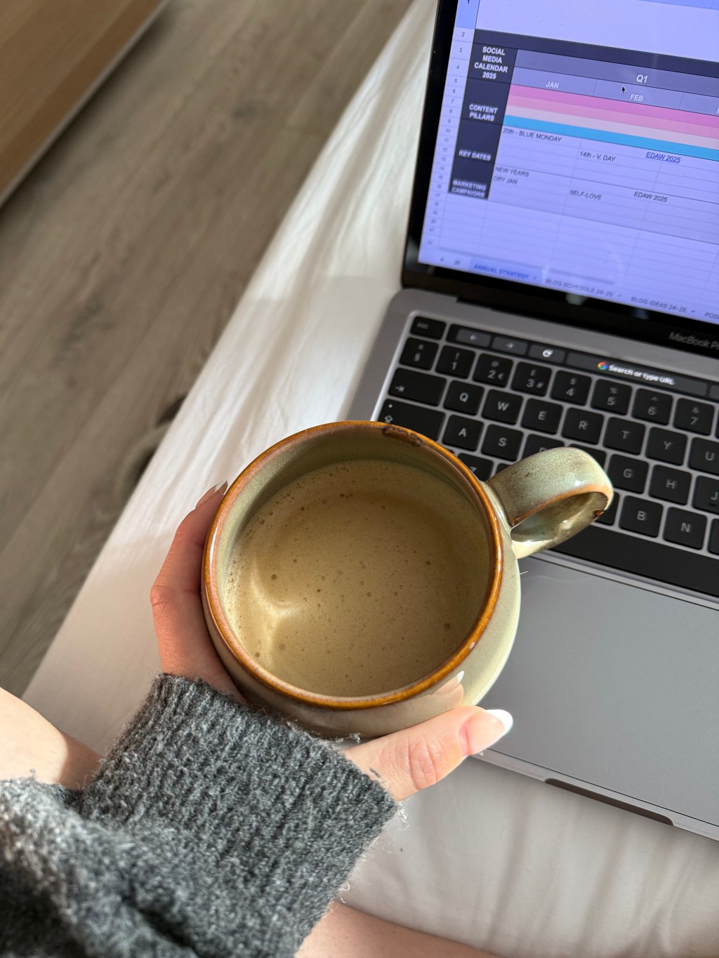 A hand holding a mug with milk coffee and a laptop with work on the screen.